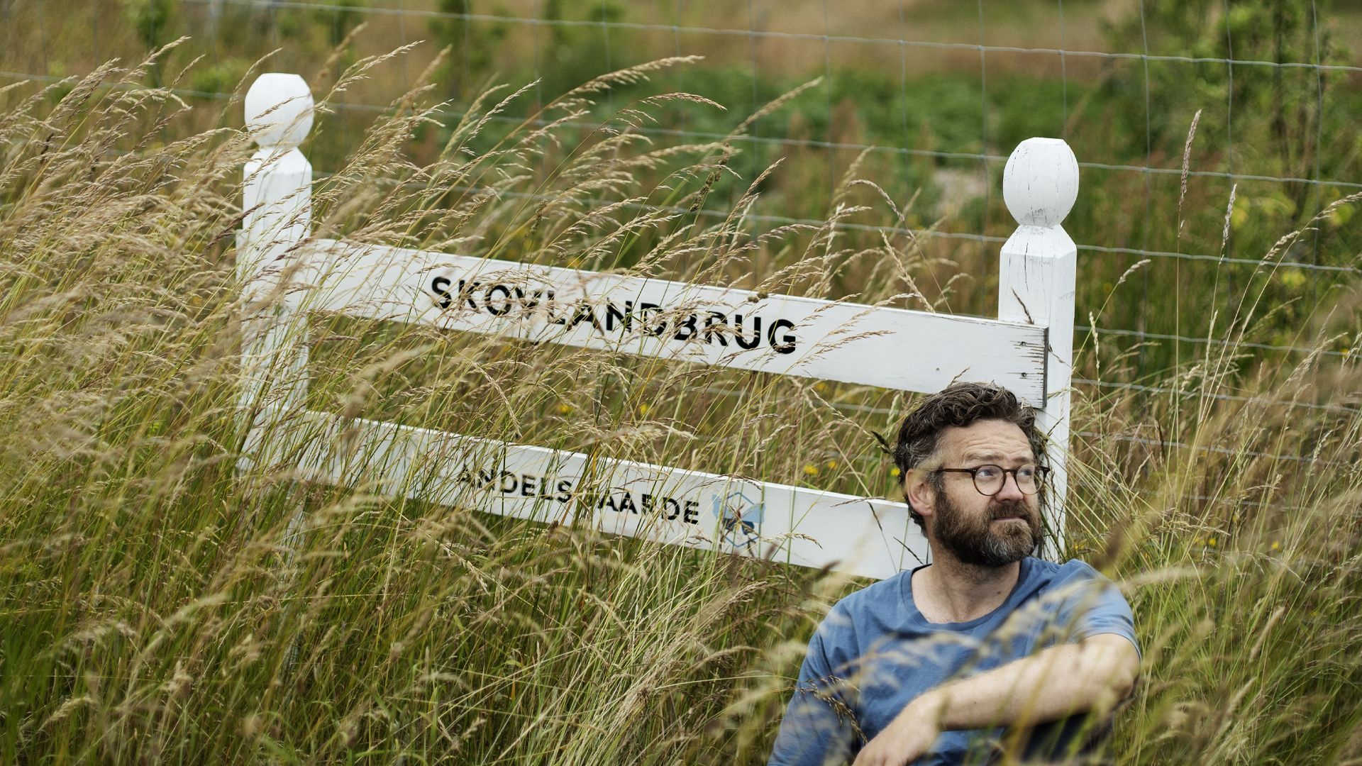 Rasmus Willig sidder i højt græs ved et skilt, hvor der står 'Skovlandbrug'